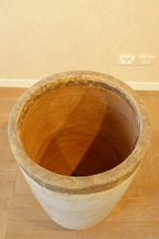 A large cylindrical concrete planter with a wide opening, shown from an elevated perspective on a wooden floor against a beige wall.